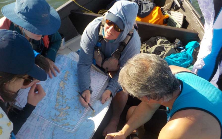A group of people sitting in a sailboat examine a map that is spread out between them. 